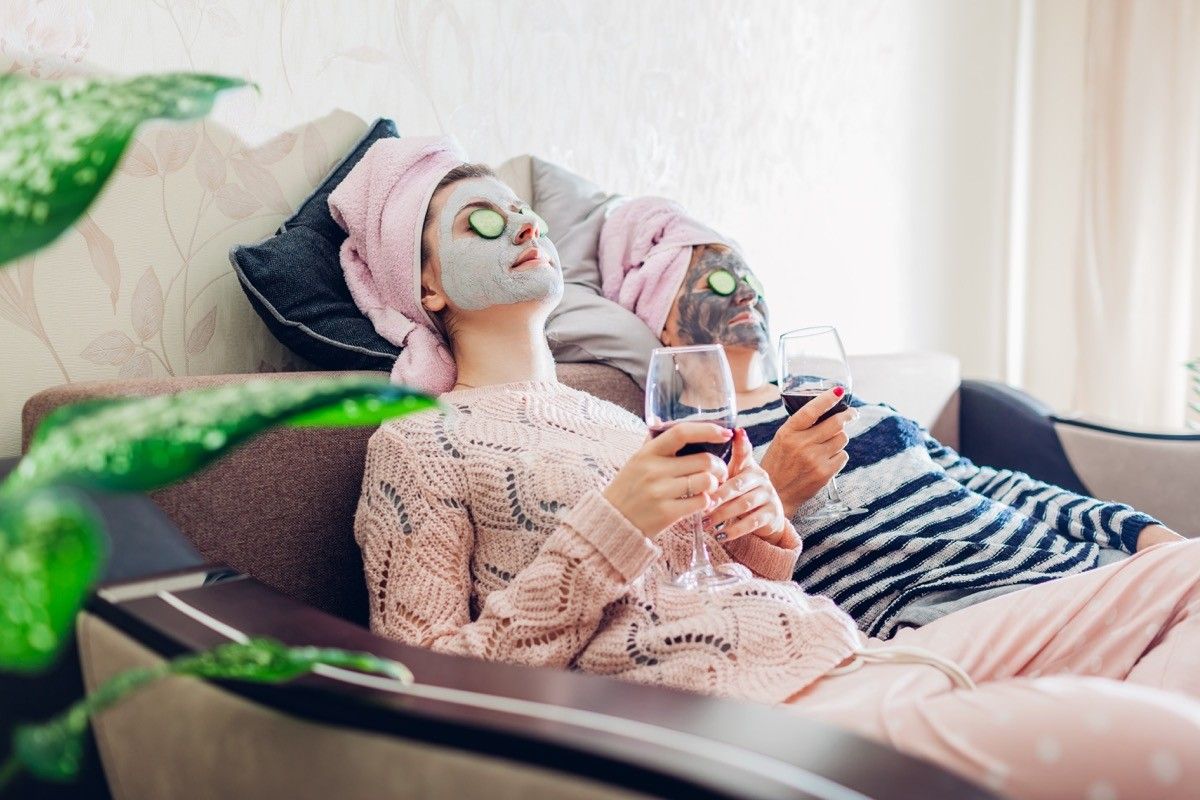 母亲和成年的女儿在眼睛上涂了面膜和黄瓜。女人坐在家里的沙发上喝着酒